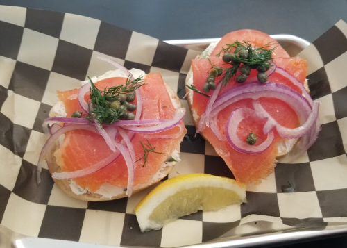 Two bagels topped with smoked salmon, cream cheese, red onion, capers, and dill, served with a lemon wedge on a checkered tray.