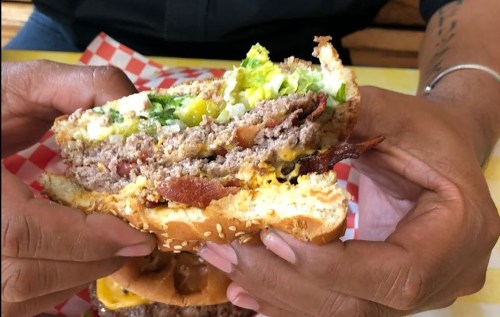 A close-up of a person holding a loaded burger with lettuce, bacon, and cheese, showcasing its layers.