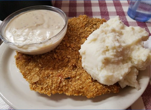 A plate featuring a large breaded fried cutlet, creamy gravy in a bowl, and a serving of mashed potatoes.