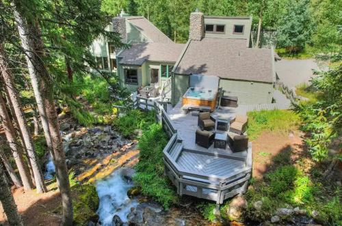A cozy cabin surrounded by trees, featuring a deck with seating and a hot tub next to a flowing stream.