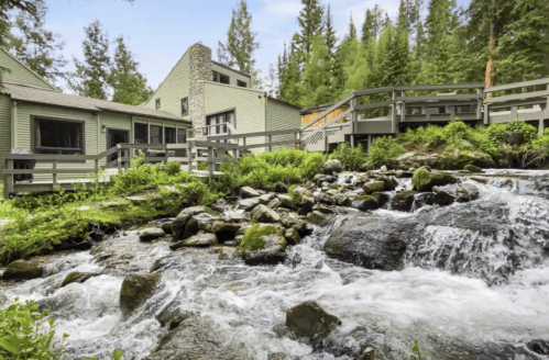 A serene landscape featuring a flowing stream beside a green cabin surrounded by trees and wooden walkways.