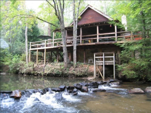 A rustic cabin by a creek, surrounded by trees, with a deck overlooking the water and a rocky shoreline.