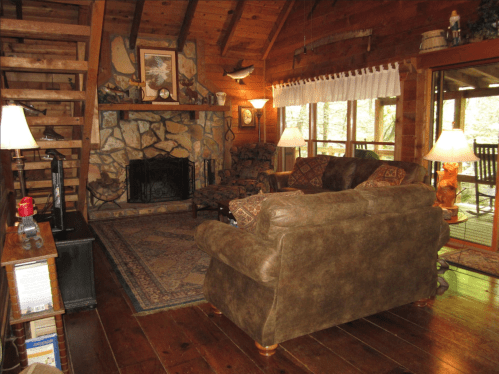 Cozy cabin interior with wooden beams, stone fireplace, comfortable seating, and warm lighting.