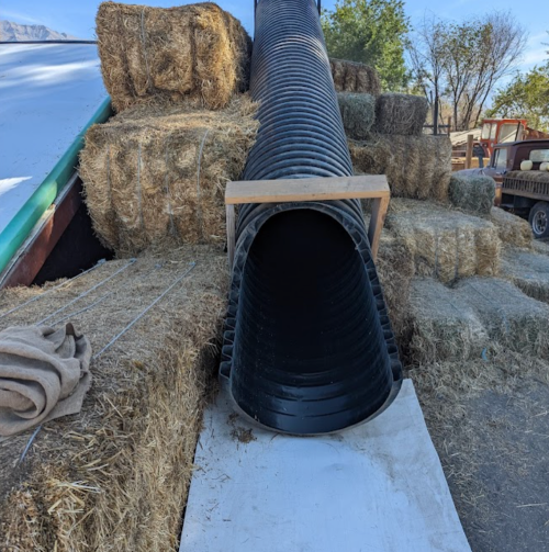 A long black tube extends from a haystack, resting on a white surface, surrounded by bales of hay and farm equipment.