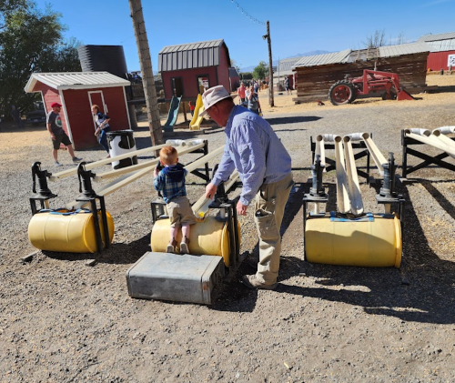 A man helps a child climb onto a yellow roller at a farm, with buildings and equipment in the background.