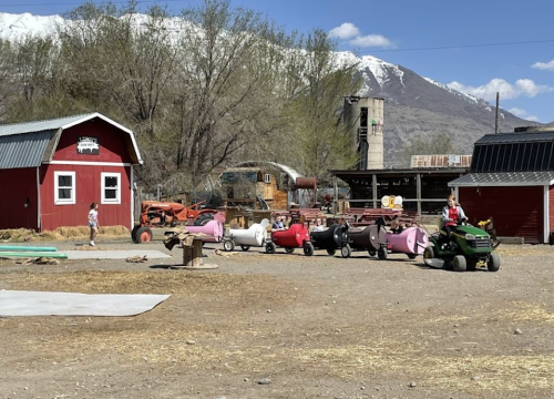 A farm scene with children riding colorful toy tractors in front of a red barn and mountains in the background.