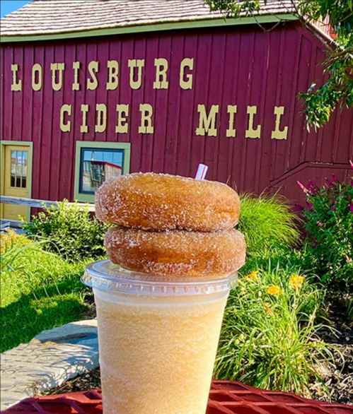 A refreshing drink topped with two cinnamon sugar donuts in front of the Louisburg Cider Mill sign.