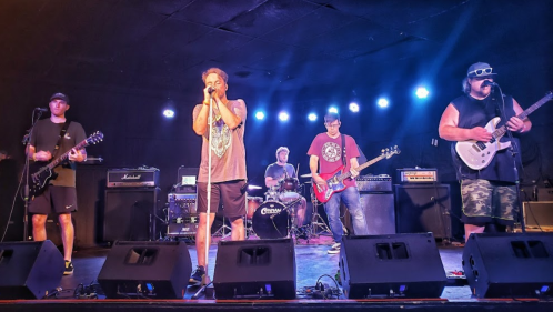 A band performs on stage with guitars and drums, under colorful stage lights in a lively venue.