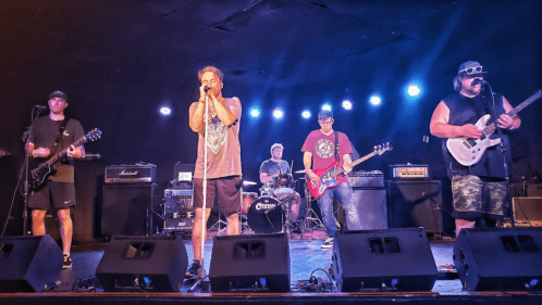 A band performs on stage with guitars and drums, under colorful stage lights in a lively venue.