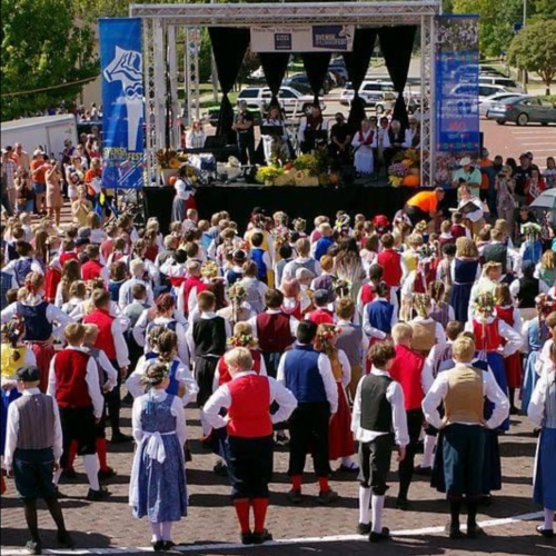 A large crowd in traditional attire gathers at an outdoor stage for a performance, with colorful decorations and a festive atmosphere.
