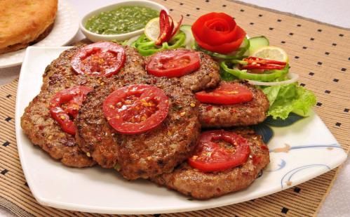 A plate of grilled patties topped with tomato slices, served with green chutney and a fresh vegetable garnish.