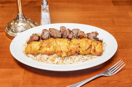 A plate of grilled chicken and beef skewers served over a bed of rice, with a fork and salt shaker in the background.