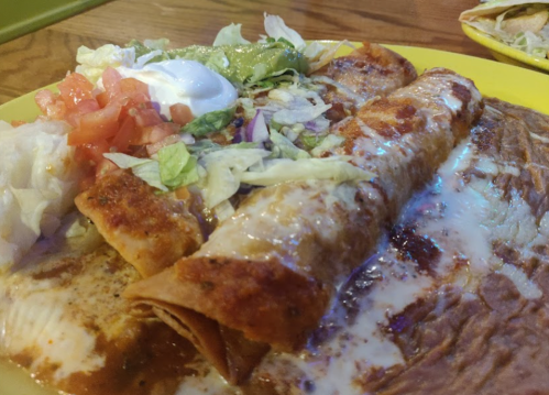 A plate of enchiladas topped with cheese, lettuce, and sour cream, served with diced tomatoes and lettuce on the side.