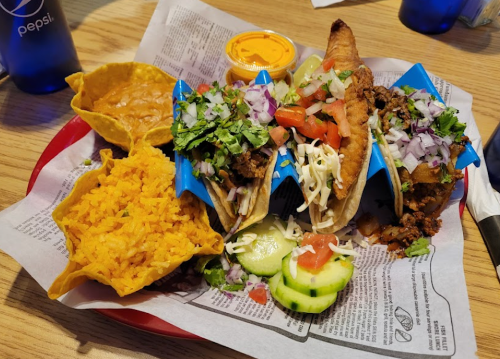 A plate of three tacos with toppings, served with rice, beans, and a side of sauce.
