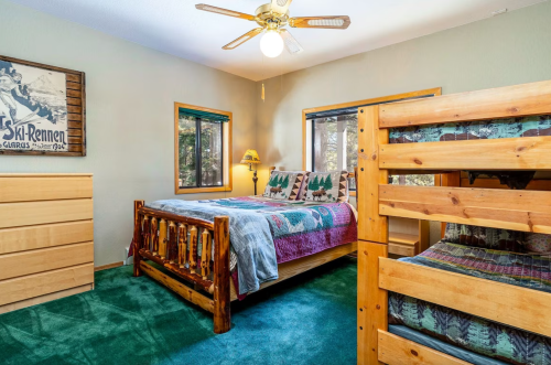 Cozy bedroom with a wooden bed, bunk bed, green carpet, and large windows, featuring rustic decor and a ceiling fan.