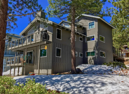 A modern gray house surrounded by trees and snow, featuring multiple windows and a deck.