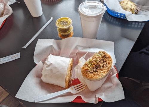 A plate with a wrapped sandwich, a burrito, and three sauce containers, alongside a cup on a table.