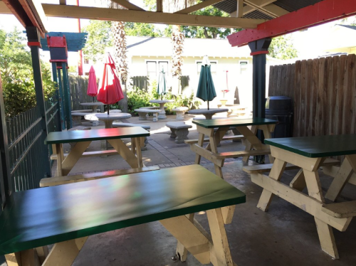 Outdoor seating area with green picnic tables, umbrellas, and a fenced patio surrounded by trees and a wooden fence.