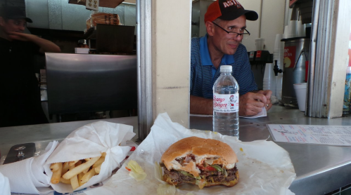 A burger and fries on a table, with a man sitting in the background at a fast-food counter. A water bottle is also visible.