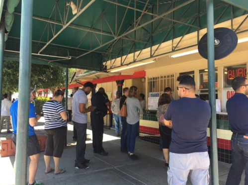 A line of people waiting outside a burger restaurant under a green awning on a sunny day.