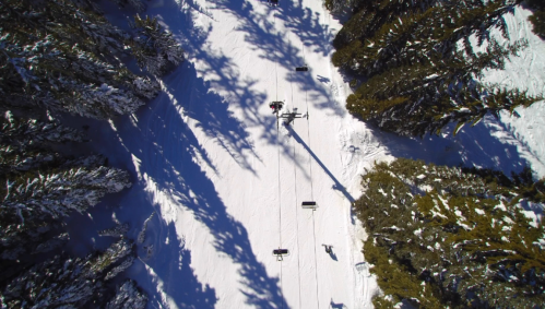 Aerial view of a snowy ski slope with ski lifts and tall trees casting long shadows.