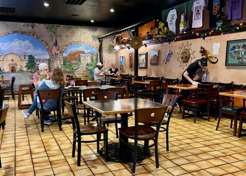 A cozy restaurant interior with wooden tables, chairs, and colorful murals on the walls, featuring a few diners.