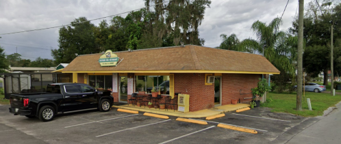 A small brick building with a yellow roof, outdoor seating, and a black truck parked in front. Palm trees nearby.