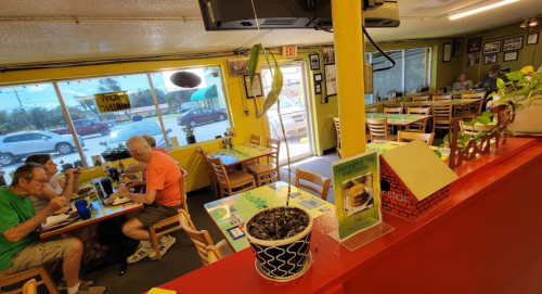 A colorful diner interior with patrons eating at tables, plants, and a view of the street outside.