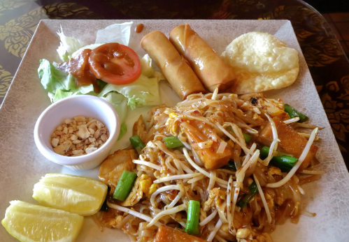 A plate of Pad Thai with spring rolls, salad, lime wedges, and a side of crushed peanuts.