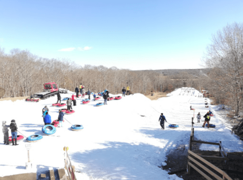 A snowy hill with people tubing, a snow groomer in the background, and trees lining the area under a clear blue sky.