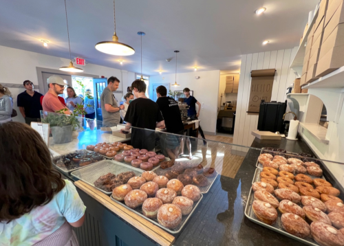 A busy bakery with customers in line, showcasing a display of various donuts in the foreground.