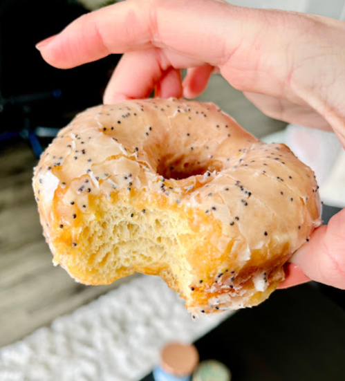 A hand holding a partially eaten glazed donut with poppy seeds on a textured surface.