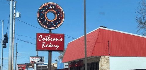 Sign for Cothran's Bakery featuring a large donut graphic against a clear blue sky. Red building in the background.
