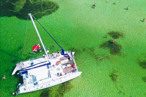 Aerial view of a catamaran anchored in clear green water, with people swimming nearby.