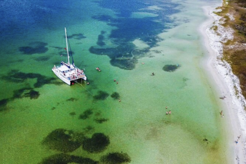 Aerial view of a sailboat anchored in clear turquoise waters, with people swimming and a sandy beach nearby.