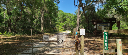 A gated entrance to a wooded area with signs for speed limit and access instructions, surrounded by lush greenery.