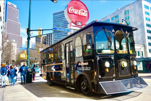 A black trolley bus parked on a city street with skyscrapers and a Coca-Cola sign in the background.