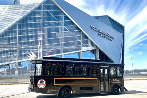 A black trolley bus parked in front of a modern stadium with a glass facade under a blue sky.