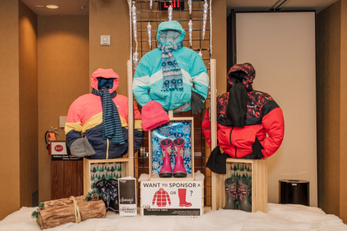 Display of colorful winter jackets, hats, and boots on a wooden stand, with a sign asking about sponsorship.