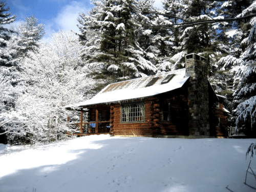 A cozy log cabin surrounded by snow-covered trees under a bright blue sky.