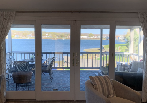 A view through glass doors of a serene waterfront scene with outdoor seating and a calm blue lake.