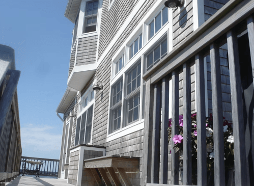 A wooden deck leads to a house with gray siding and large windows, featuring flowers in a planter.