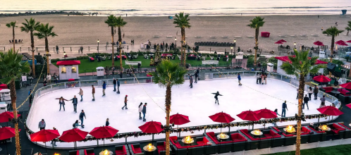 An outdoor ice skating rink by the beach, surrounded by palm trees and red umbrellas, with people skating and enjoying the scene.