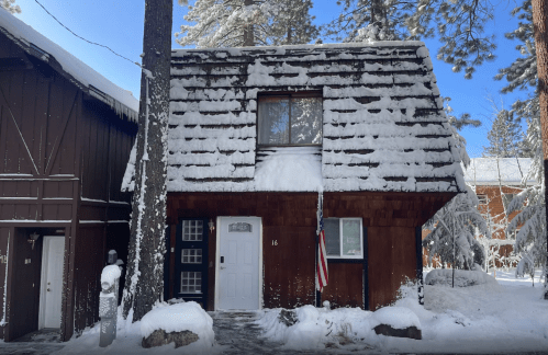 A snow-covered cabin with a steep roof, surrounded by trees, featuring a front door and an American flag.