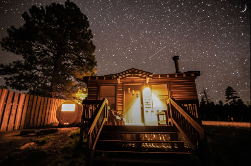 A cozy cabin illuminated at night, surrounded by a starry sky and trees, with a faint crescent moon above.