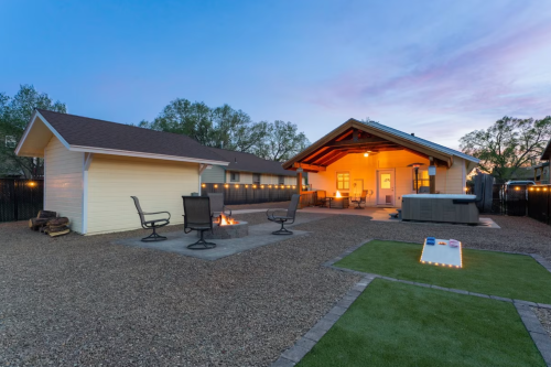 A cozy backyard with a fire pit, seating area, hot tub, and a lawn, illuminated at dusk.