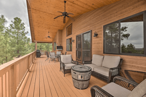 A wooden deck with seating, a table, and fans, surrounded by trees under a cloudy sky.