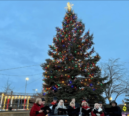 A choir sings in front of a brightly lit Christmas tree against a twilight sky.