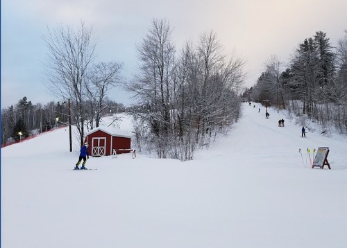 A snowy ski slope with a red barn, trees, and skiers on the hill under a cloudy sky.