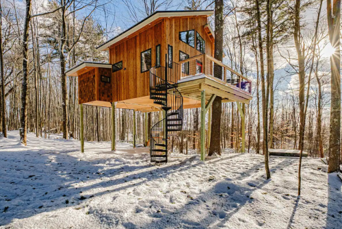 A wooden treehouse on stilts surrounded by snow-covered trees, featuring a spiral staircase and large windows.